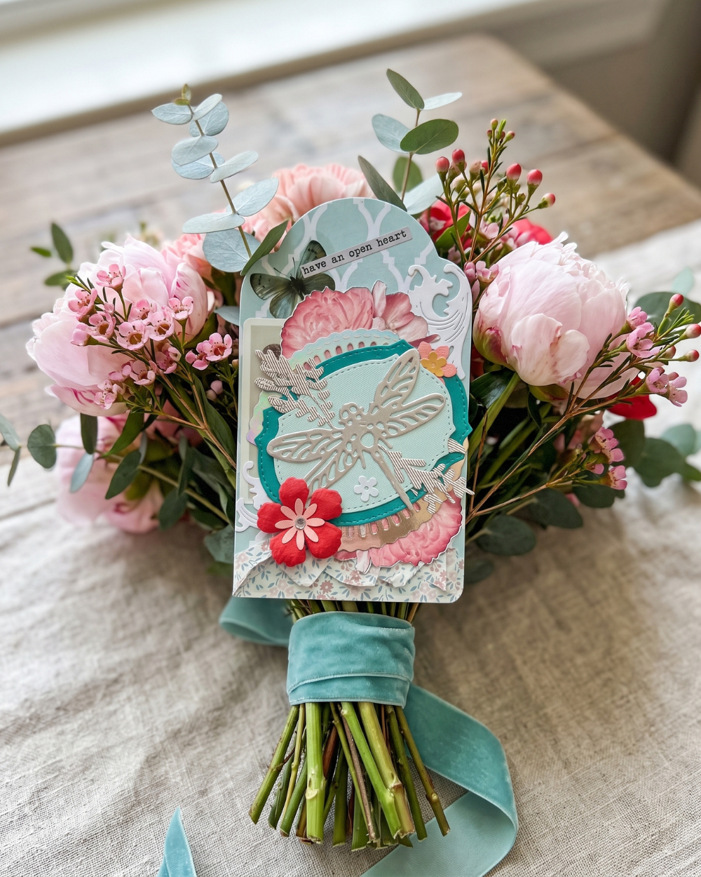 A styled bouquet of pink peonies, red filler flowers, green eucalyptus stems, and a teal ribbon wrap, with a decorative layered paper dragonfly tag attached to the center.