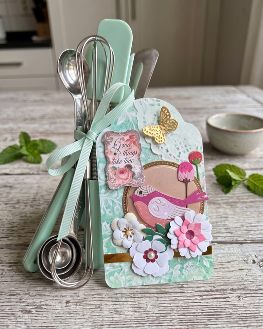 A decorative kitchen utensil set displayed on a rustic wooden table, featuring mint green silicone spatulas and measuring cups tied with a mint green ribbon, alongside a pink bird embellishment, flowers, and a gold butterfly on the front tag.