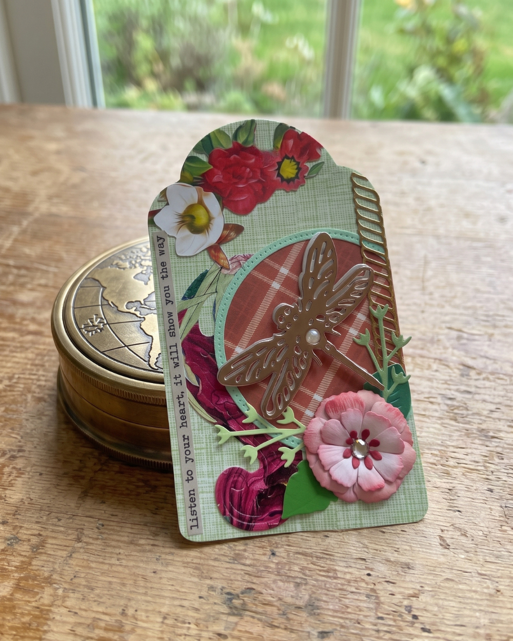 A handmade layered paper card or pin-back art piece with floral designs, a dragonfly embellishment, and a pink flower, displayed on a wooden table next to a round brass tin.