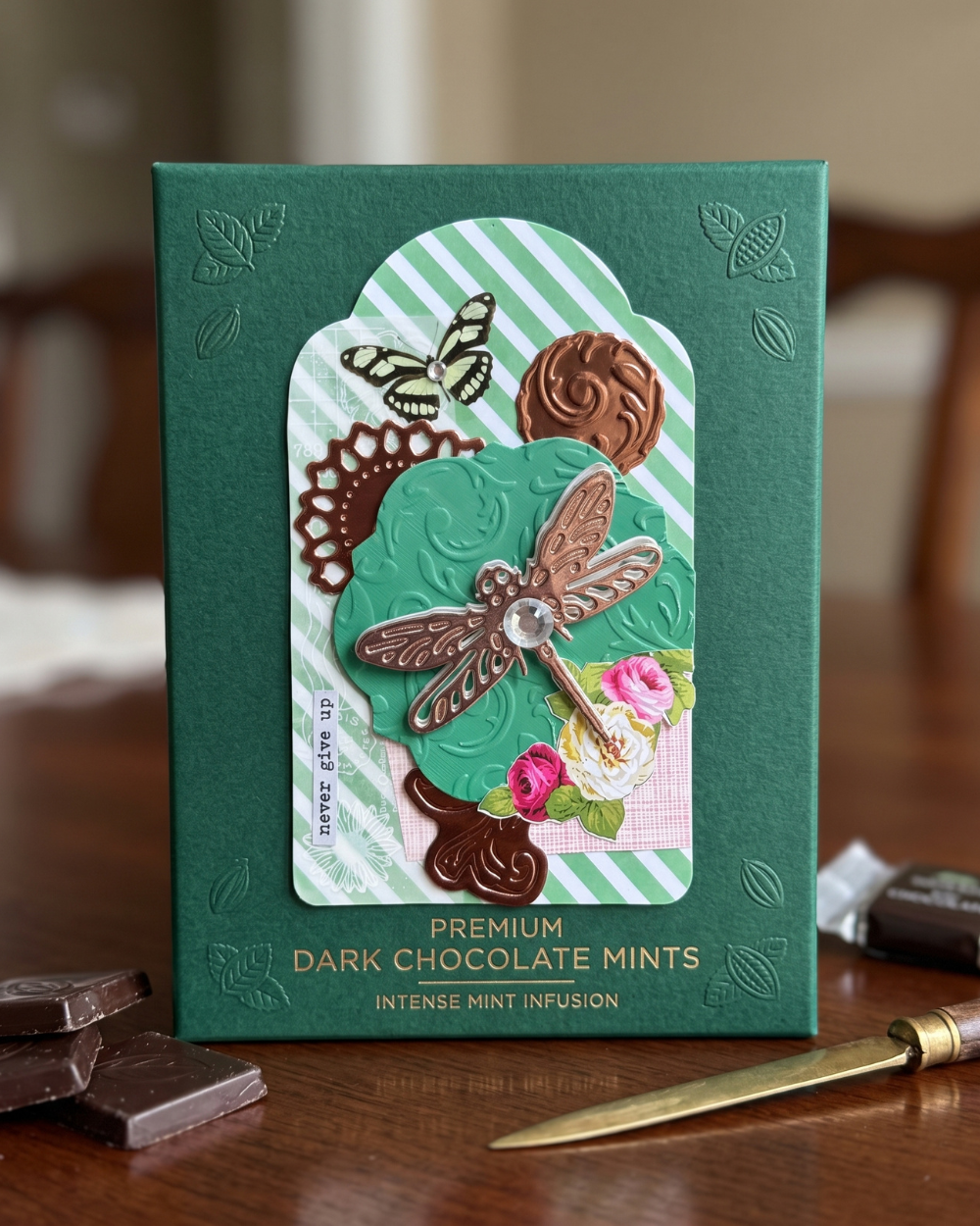 A green rectangular box of premium dark chocolate mints sits on a wooden table. The front of the box features an ornate layered design with a dragonfly, flowers, gears, and a striped background, along with the text 'PREMIUM DARK CHOCOLATE MINTS' and 'INTENSE MINT INFUSION.