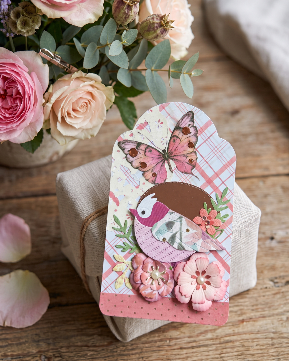 A decorative handmade paper card featuring a pink butterfly and bird design, styled on a rustic wooden surface next to a vase of pink and peach roses.