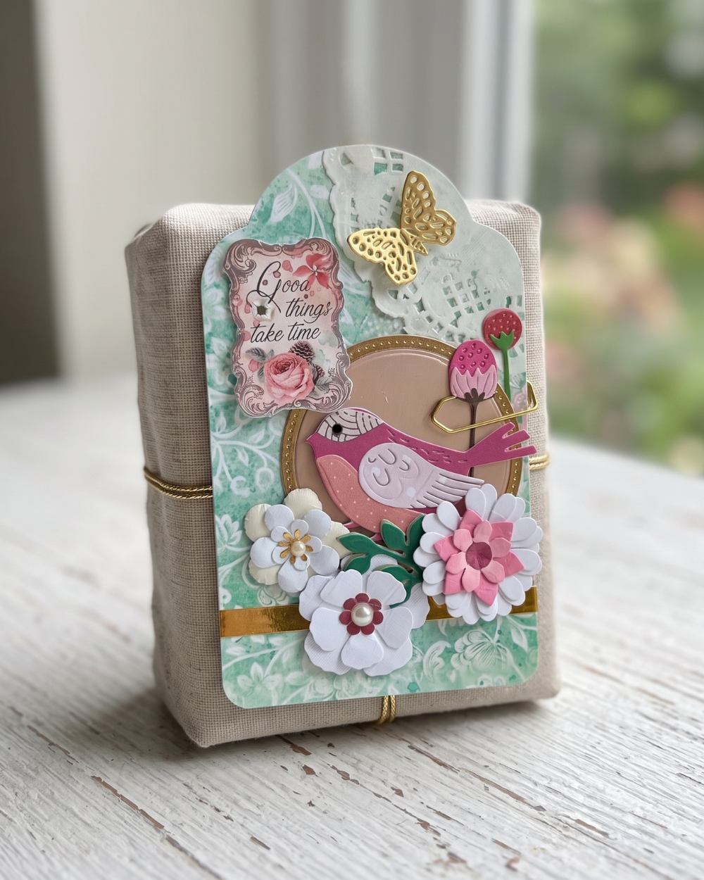 A handmade decorative gift box wrapped in beige linen, adorned with a layered paper label featuring a pink bird, white and pink flowers, a gold butterfly, and a tag that reads 'Give me time take time'.