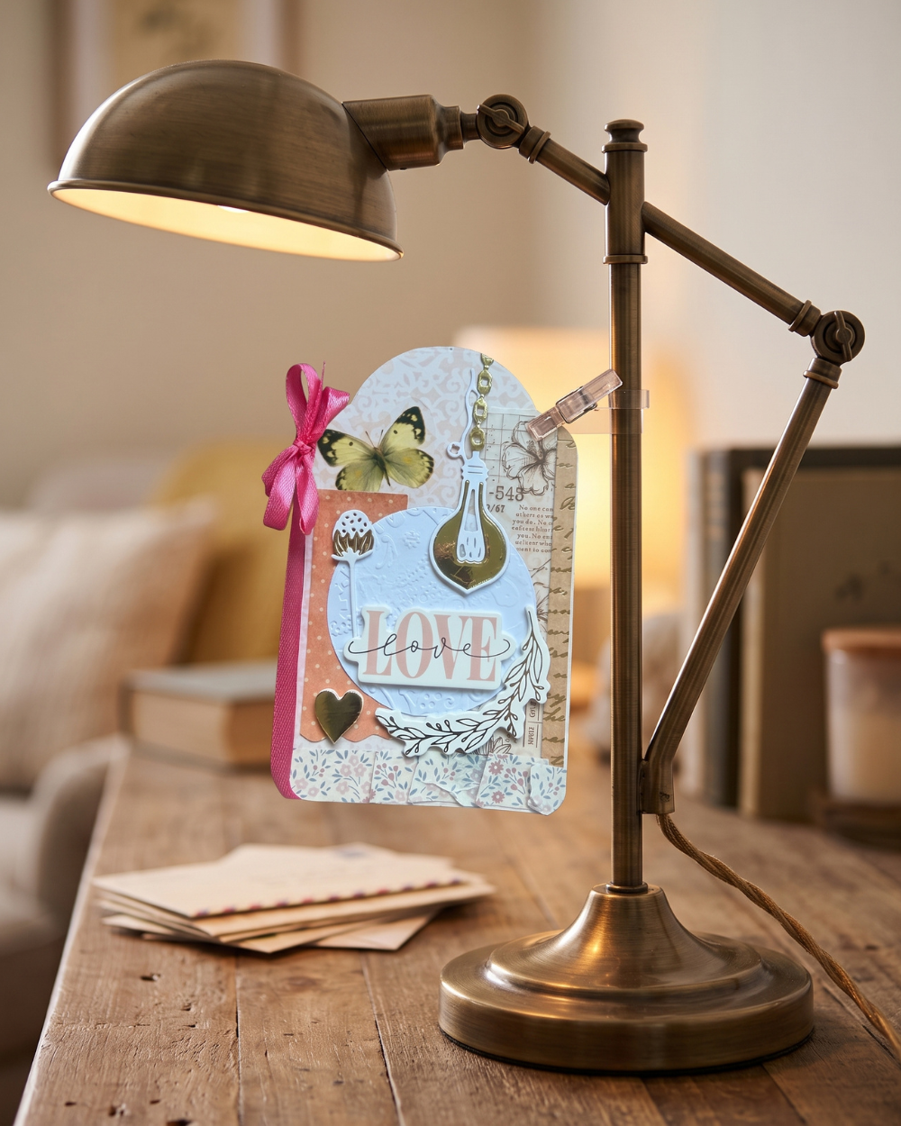 A vintage-style adjustable desk lamp with a warm brass finish sits on a wooden table in a cozy room setting. A decorative handmade card with a pink ribbon bow and butterfly clip is hanging from the lamp arm.
