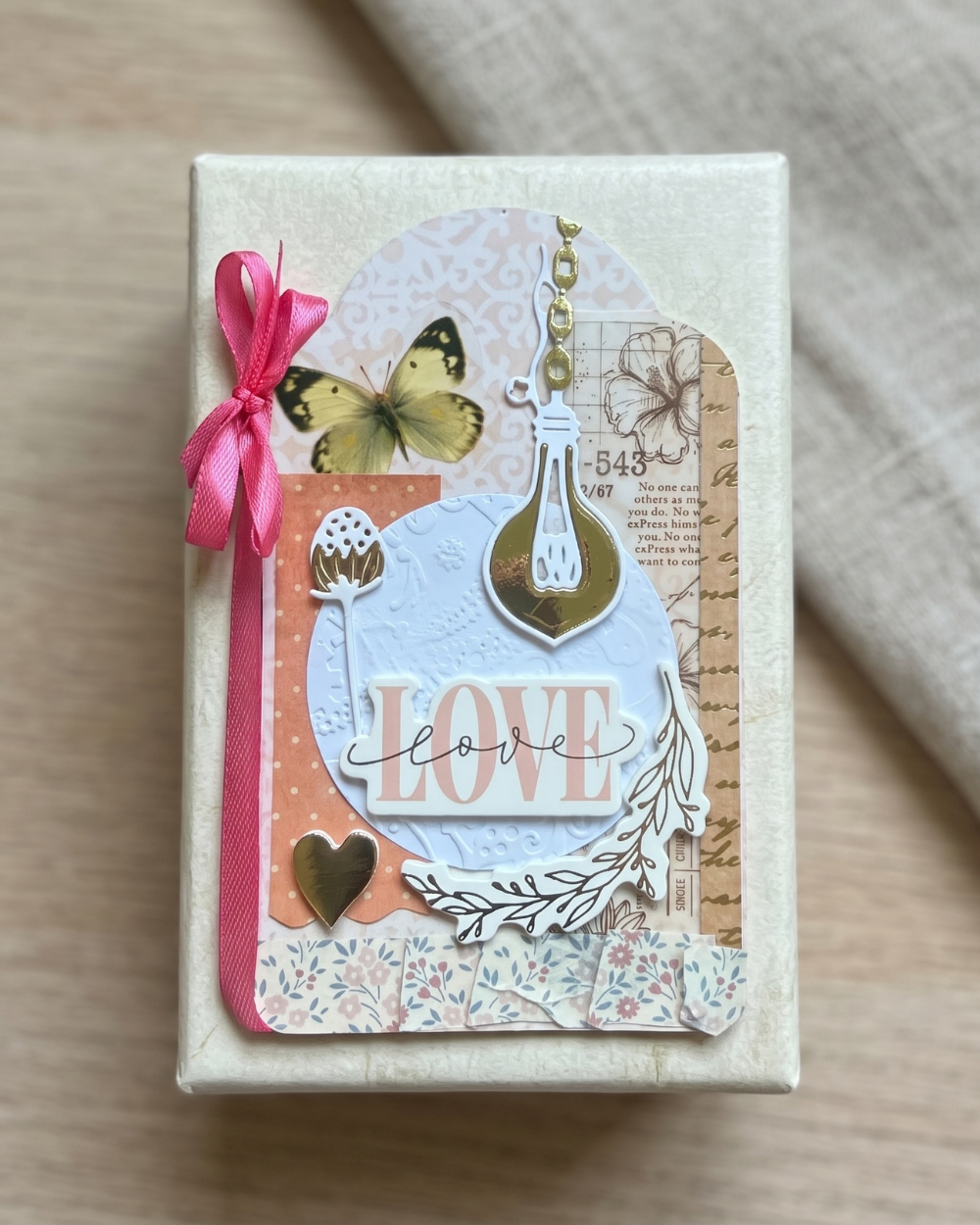 A square cream-colored gift box wrapped in textured paper, decorated with layered vintage-style paper cutouts, a pink ribbon bow, a butterfly, a lightbulb charm, and the word 'LOVE' in script, set on a light wood surface with a beige fabric background.