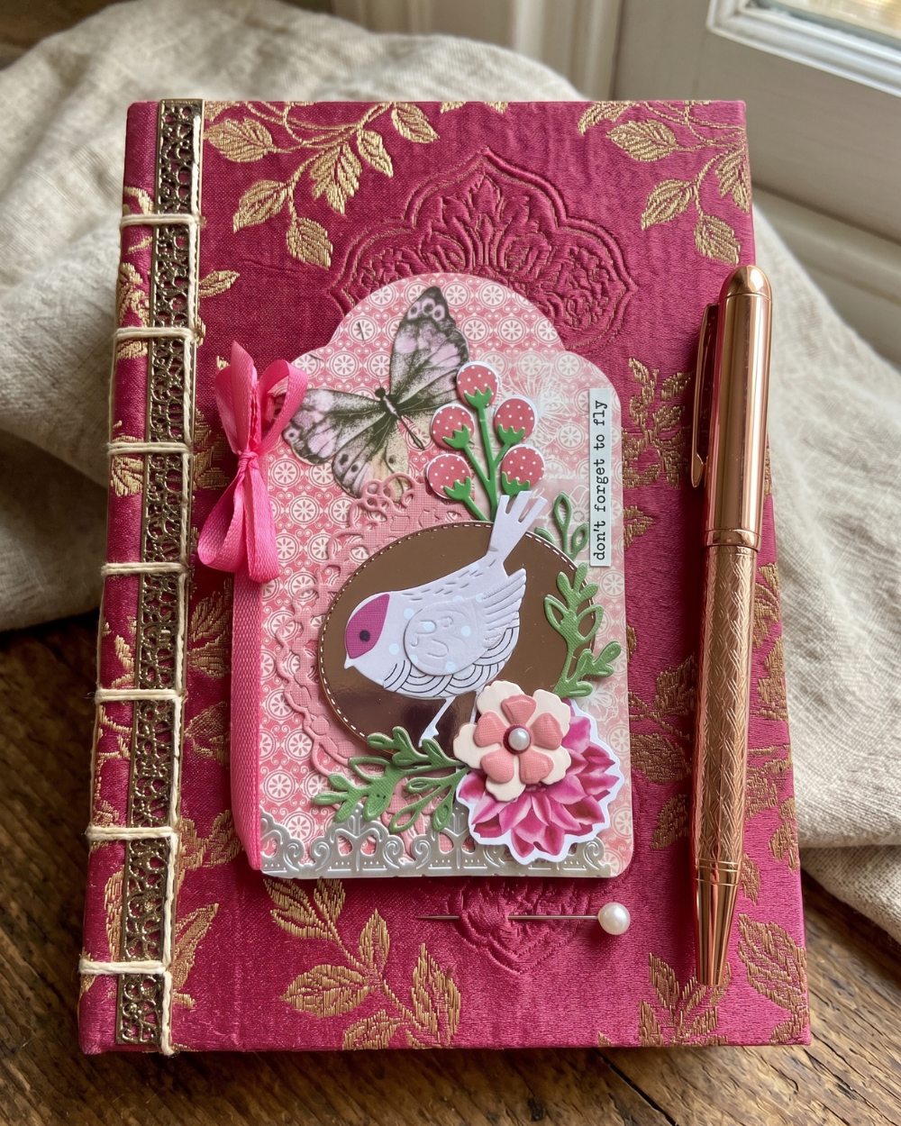 A decorative pink embossed journal with gold floral detailing, a bird and butterfly embellishment on the cover, a pink ribbon bow, a pen, and a small pearl pin, photographed on a wooden surface with draped fabric in the background.