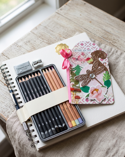 A styled flat-lay of a metal pencil case set on a rustic wooden table. The set includes black and colored pencils, a white eraser, a pink ribbon, and a decorative dragonfly-themed notebook cover.