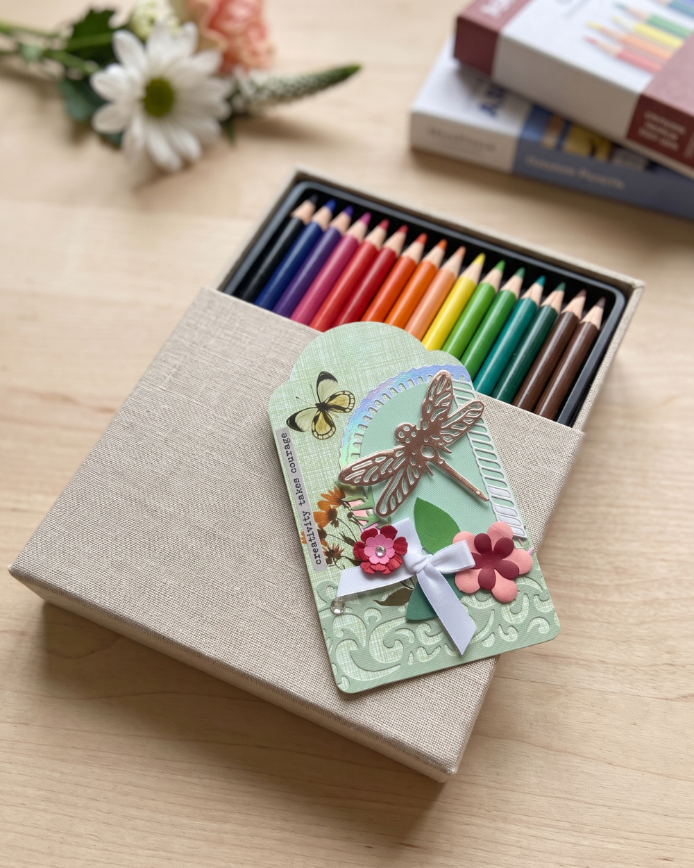 A gift box of colored pencils with a decorative paper tag attached, placed on a light wood surface. The box is open to show multiple rows of pastel and bright colored pencils, and the tag features a dragonfly, flowers, and butterflies.