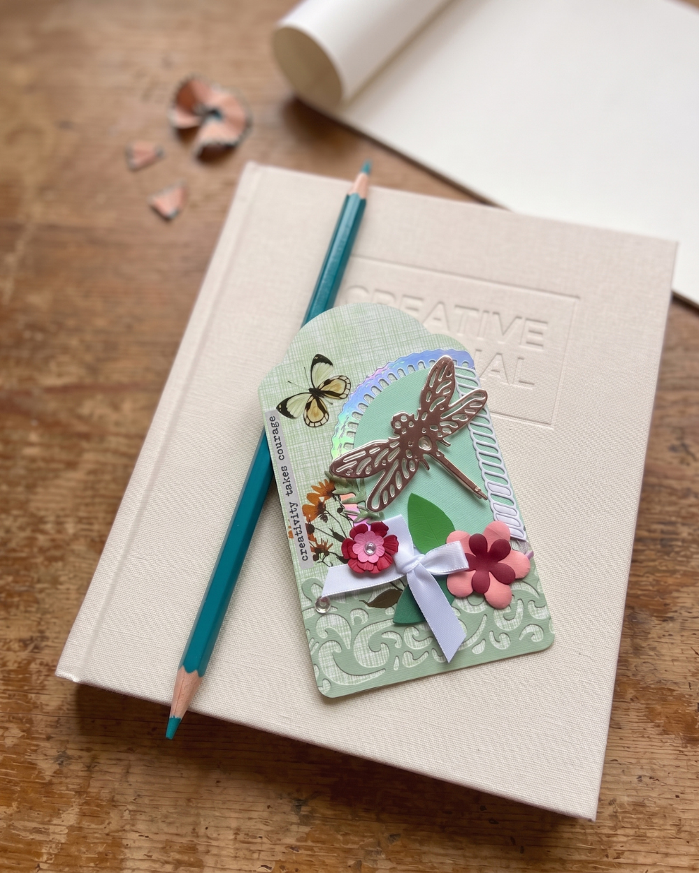 A decorative handmade paper craft tag with a dragonfly, flowers, and butterfly embellishments, styled on top of a cream-colored sketchbook with a teal pencil on a wooden table.