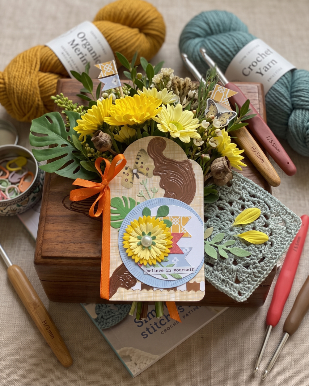 A styled flat-lay of a handmade crochet gift set on a beige fabric background, featuring a wooden box filled with yellow flowers, a decorative card, crochet hooks, yarn skeins, a crochet square, and a knitting needle.
