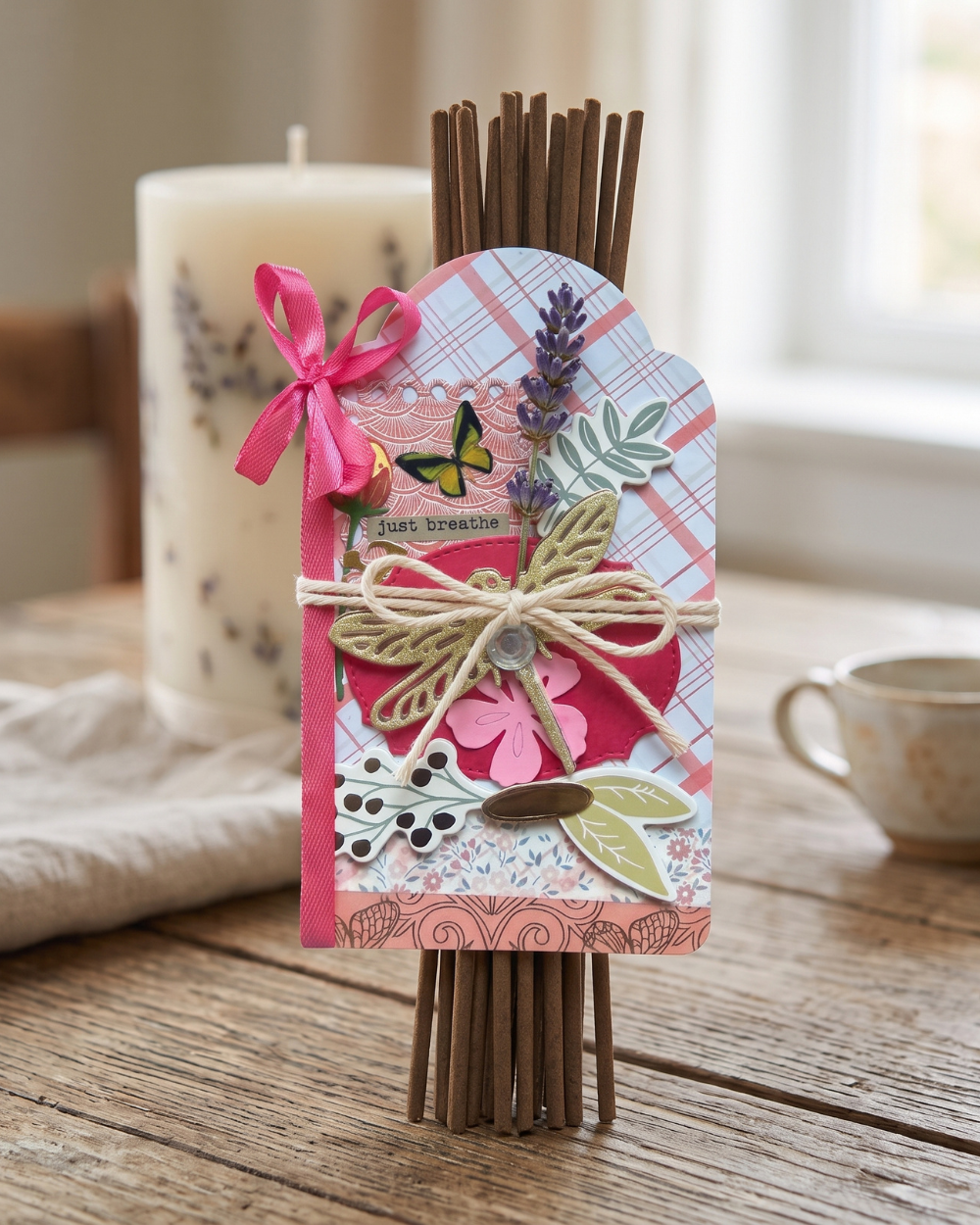 A decorative bundle of incense sticks tied with twine and a pink paper tag, styled on a rustic wooden table with a candle and teacup in the background.