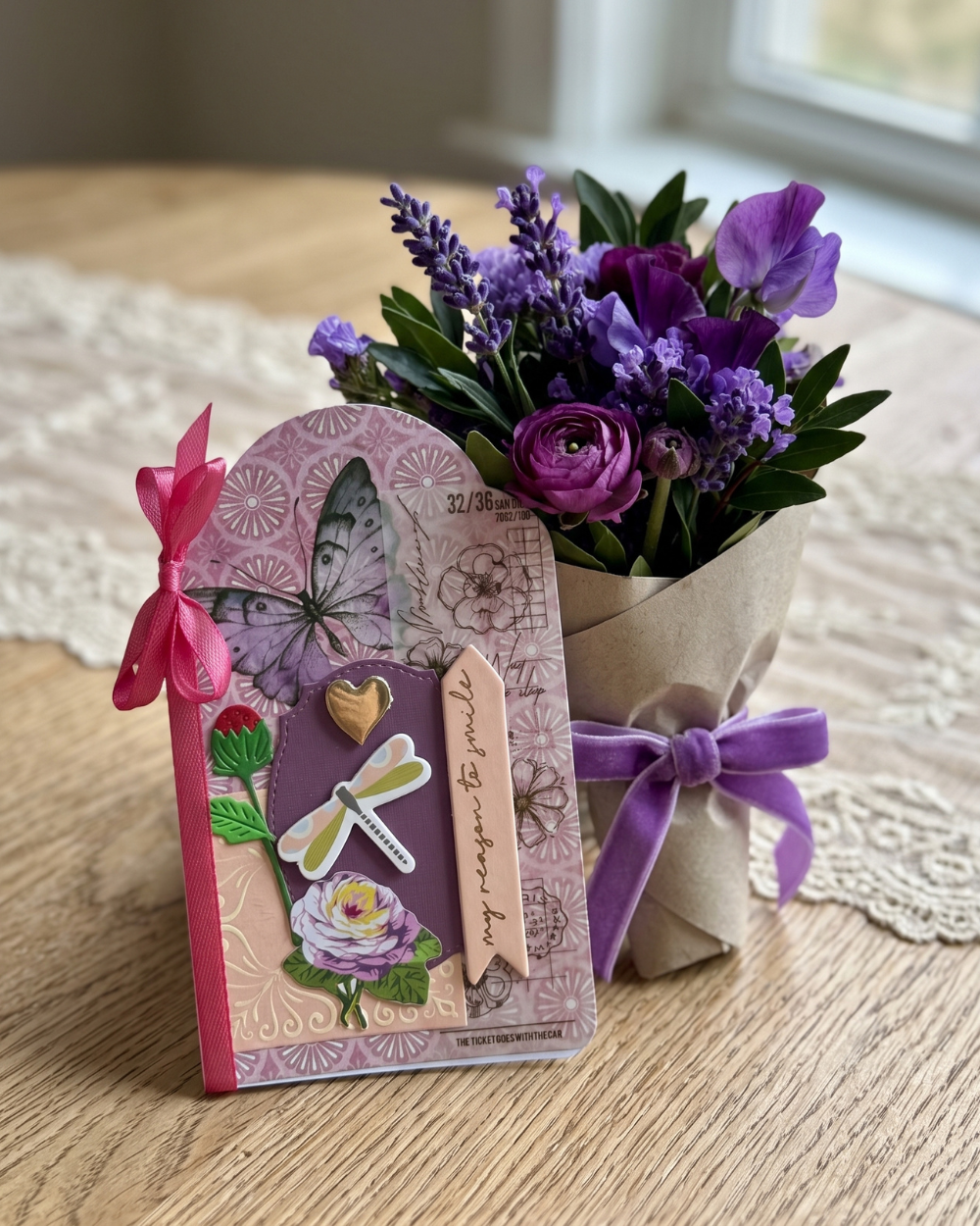 A handmade paper card with a butterfly and dragonfly design stands next to a small bouquet of purple flowers wrapped in kraft paper with a purple ribbon, set on a wooden table.