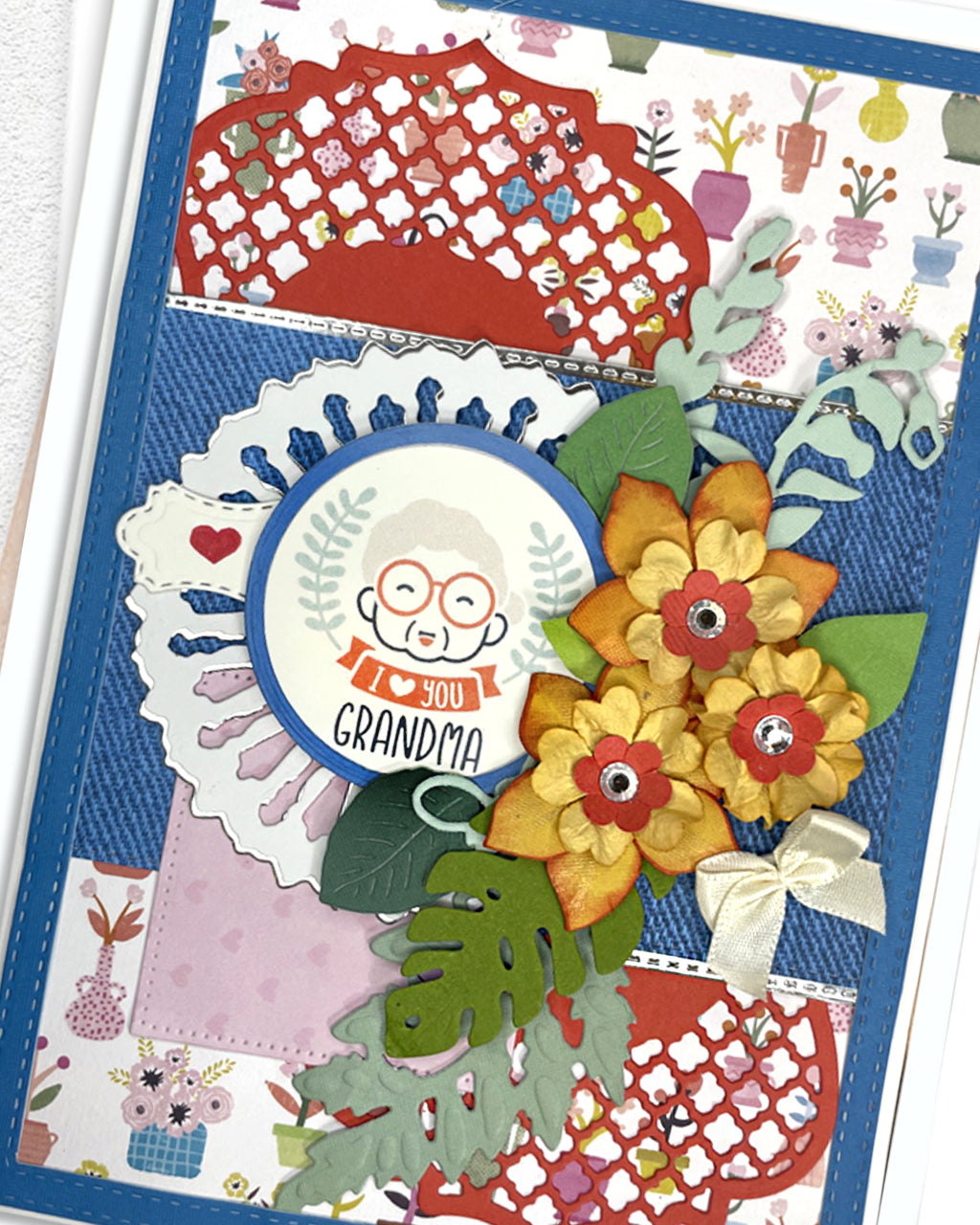Close-up of a handmade grandma card showing three layered yellow paper daisy flowers with red gem centres, large green tropical leaves, and a circular "To my Grandma" illustrated sentiment tag on a white scalloped frame. Vibrant patchwork patterned background in red, teal and cobalt blue. Handmade greeting card, New Zealand.

