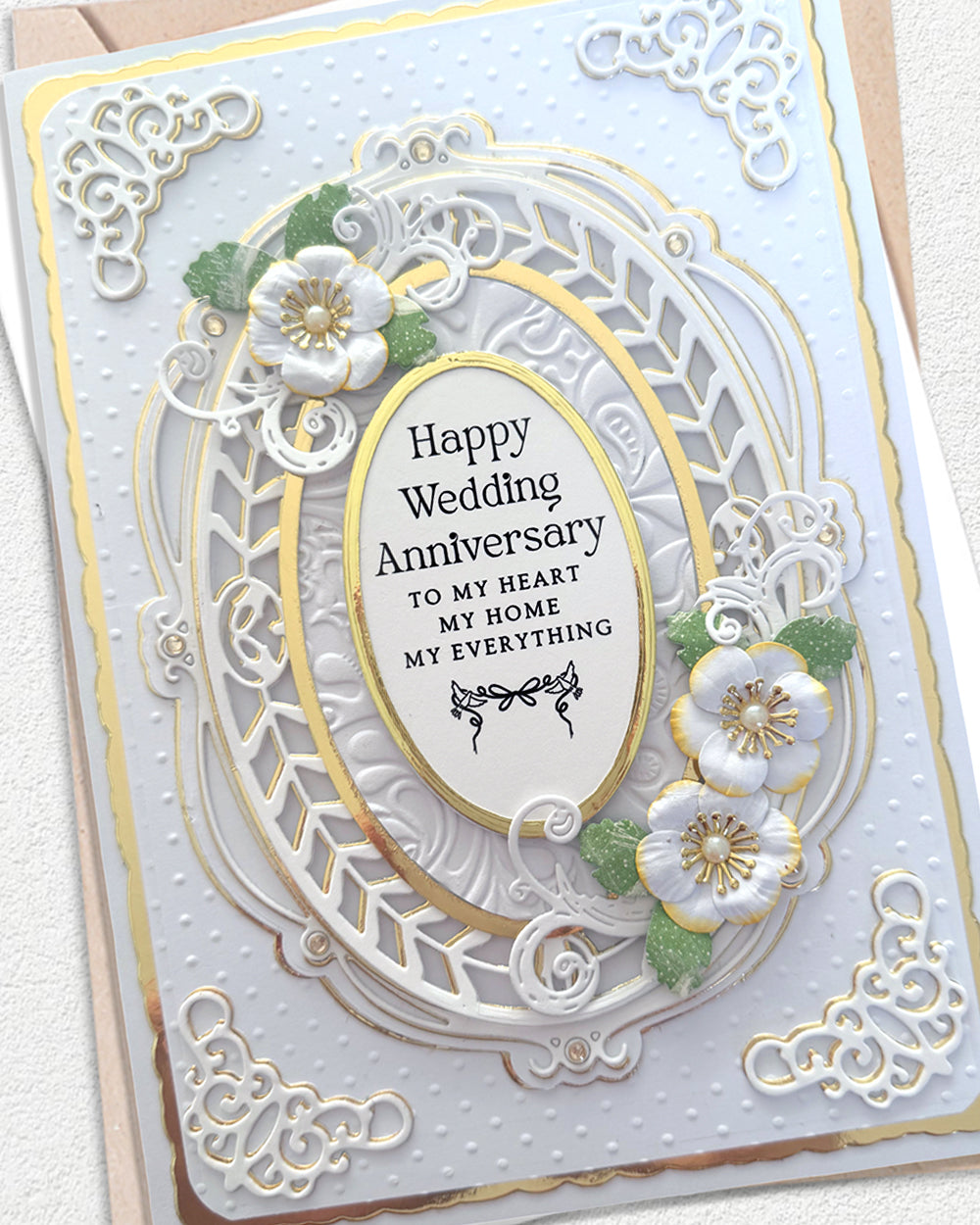 A handmade wedding anniversary card with intricate white laser-cut floral and scroll designs, gold foil accents, and 3D white flowers with green leaves. The central oval reads 'Happy Wedding Anniversary TO MY HEART MY HOME MY EVERYTHING'.