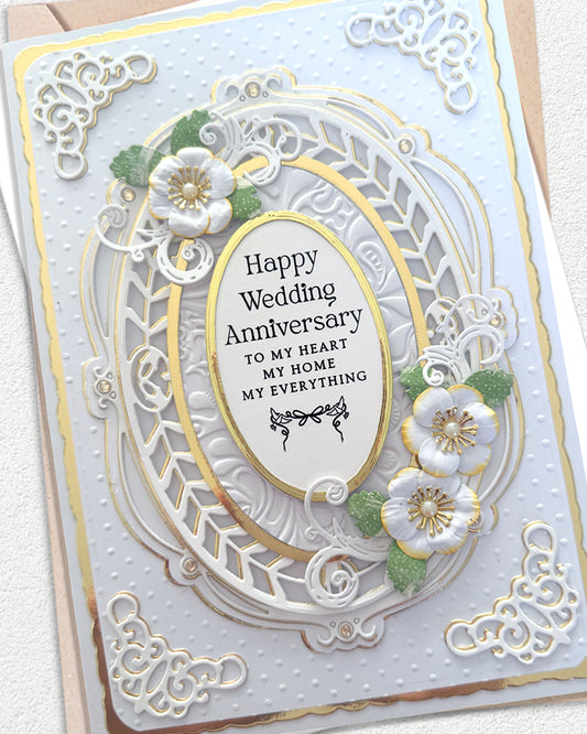 A handmade wedding anniversary card with intricate white laser-cut floral and scroll designs, gold foil accents, and 3D white flowers with green leaves. The central oval reads 'Happy Wedding Anniversary TO MY HEART MY HOME MY EVERYTHING'.