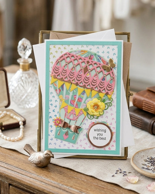 A handmade greeting card featuring a colorful hot air balloon design, displayed in a gold frame on a rustic wooden table with decorative props like a glass decanter, pearl necklace, and small bird figurine.