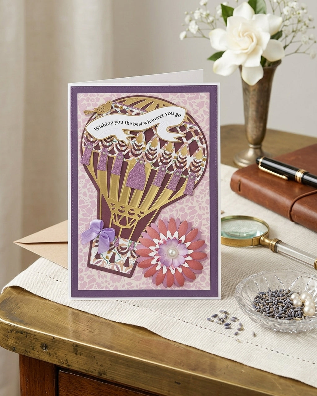 A greeting card featuring a purple and gold hot air balloon design with a pink flower and a ribbon banner reading 'Wishing you the best wherever you go,' displayed on a wooden table with a vase of white flowers and other decorative items in the background.