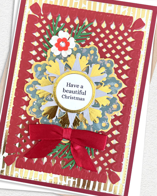 A handmade layered Christmas greeting card featuring a red scalloped border, a large red bow, a gold and blue floral mandala, green pine branches, and a white flower with a red center. The center contains a gold oval with the text 'Have a beautiful Christmas'.