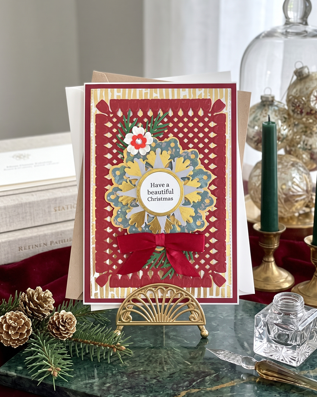 A handmade Christmas greeting card displayed on a small gold stand on a green marble surface, surrounded by holiday props including pinecones, books, a candle, and glassware. The card features a red and gold geometric border, a blue and yellow floral mandala, and a central white circle that reads 'Have a beautiful Christmas'.