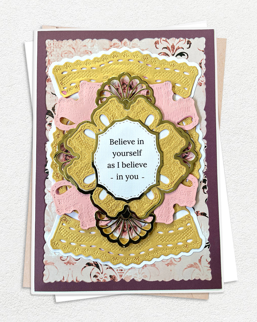 Handmade luxury encouragement card on a deep plum cardstock base, layered with blush pink and antique gold die-cut panels in graduating scalloped and ornate shapes. A large gold embossed decorative frame with fan shell detail at the base centres a white sentiment panel reading "Believe in yourself as I believe in you" in fine print. Floral patterned paper peeks through the layered die-cuts adding delicate detail. White envelope visible beneath. Handmade luxury card made in New Zealand.
