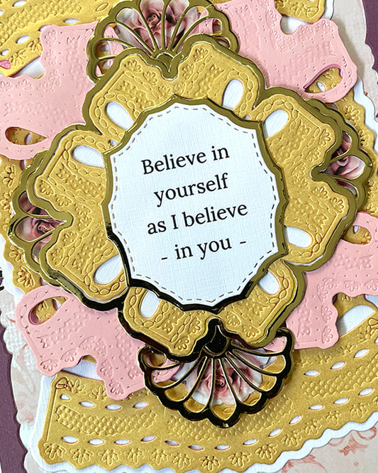 Close-up of a handmade luxury card showing an ornate antique gold embossed die-cut frame with decorative fan shell base, centring a white sentiment panel reading "Believe in yourself as I believe in you." Layered blush pink and gold die-cut panels visible behind. Handmade greeting card made in New Zealand.