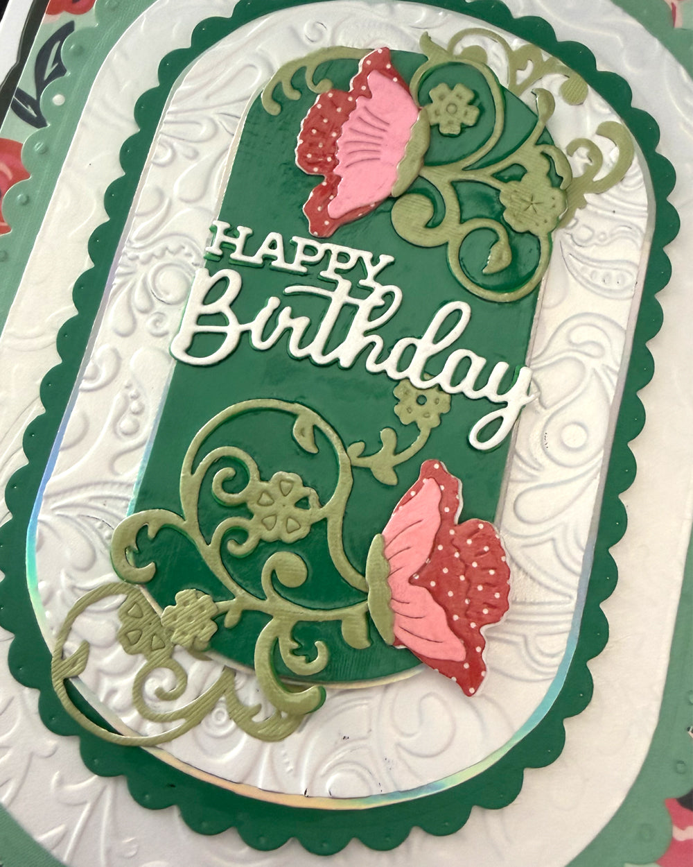 Close-up of "Happy Birthday" in white script on emerald card, with hand-finished gold floral vine and pink poppy bloom die-cuts
