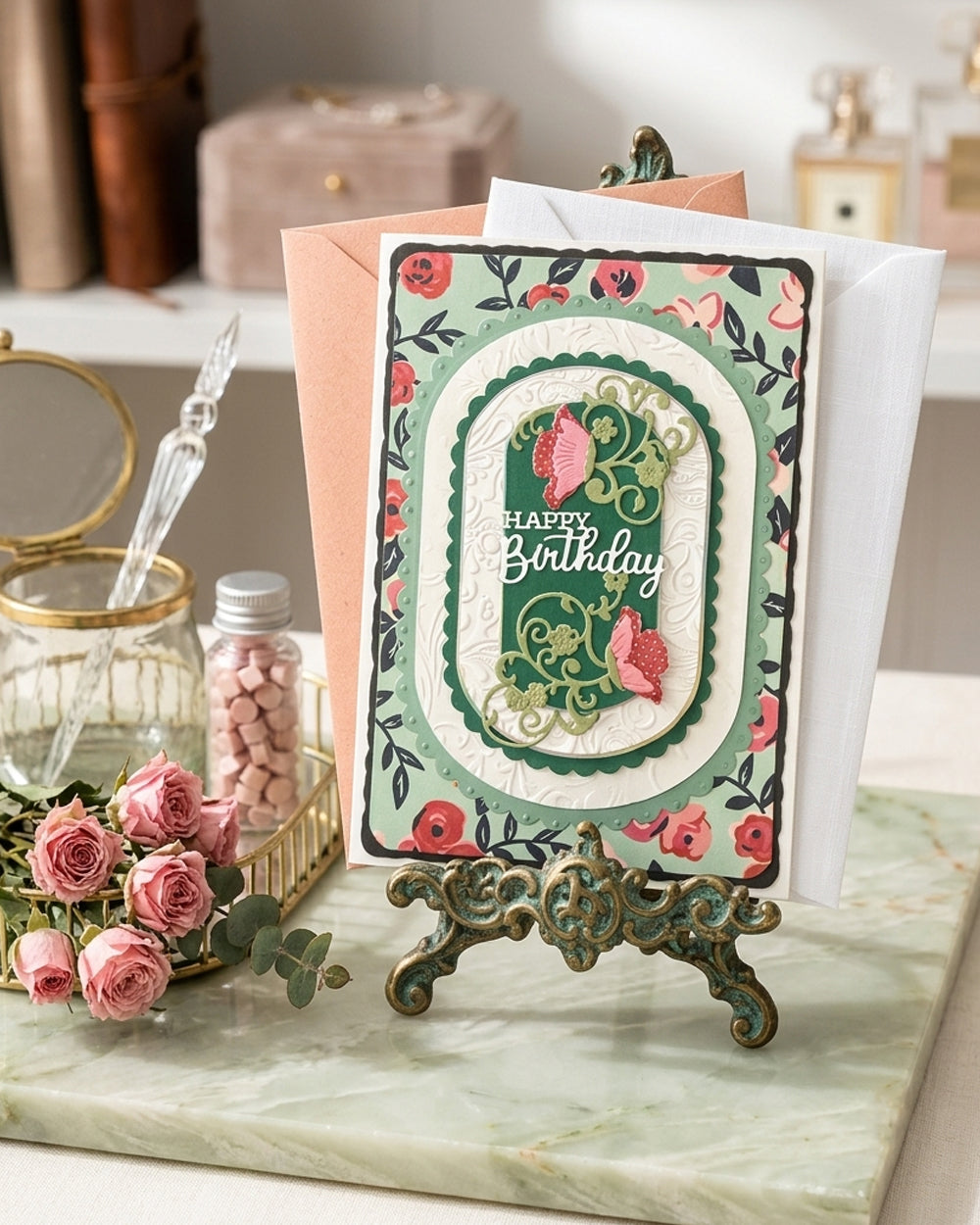 Happy Birthday card with emerald floral design displayed on a stand beside roses and a vase, styled on a green tabletop. Made in NZ by Del Cards.
