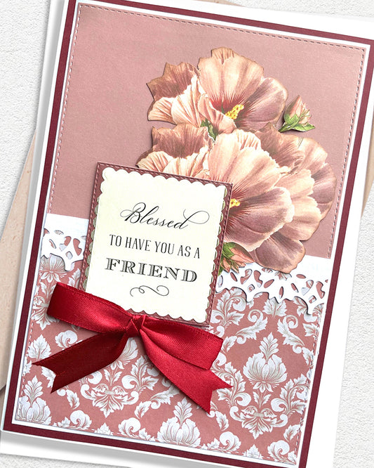 Close-up of a handmade luxury friendship card showing large vintage peach and blush paper peonies against a dusty rose background, with a cream scalloped sentiment tag reading "Blessed to have you as a Friend" and the top of a deep red satin ribbon bow. Made in NZ by Del Cards.