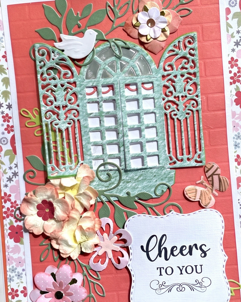 Full front view of a handmade coral and teal greeting card featuring an ornate filigree garden gate die-cut, small perched bird, hand-applied paper florals in pink and cream, ditsy floral border paper and a "Cheers TO YOU" sentiment tag. A joyful, occasion-ready handmade card made in New Zealand — perfect for celebrations, congratulations or birthdays.