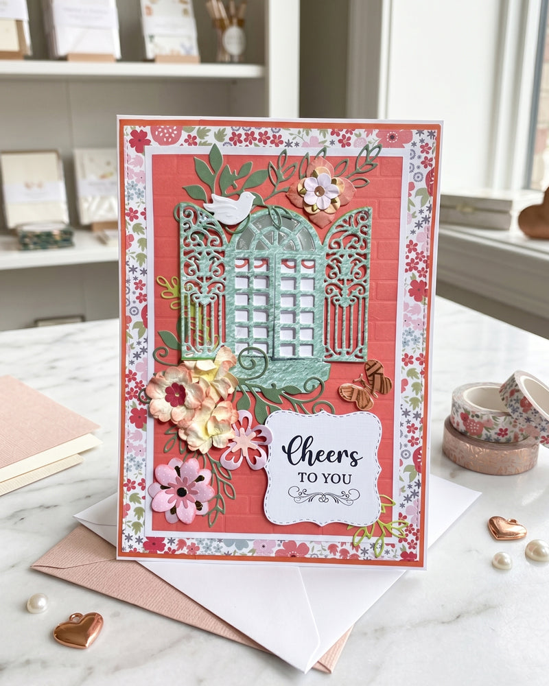 Handmade "Cheers to you" card displayed on a light surface in a soft styled setting with props alongside. Warm coral and teal tones with hand-applied paper florals make this a vibrant, celebratory handmade greeting card made in New Zealand by Del Cards.