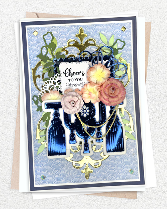 Handmade luxury celebration card on a soft blue-grey wave-textured background with a navy blue cardstock mat. An ornate gold filigree frame arches above, with green leaf sprays adding freshness. Hand-applied paper roses in blush, peach and dusty rose cluster at the centre. Sentiment reads "Cheers to You" in elegant script on a white tag. Handmade in NZ by Del Cards.