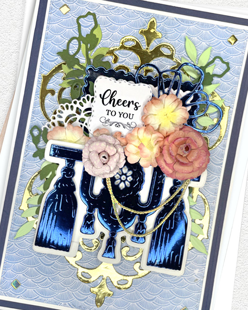 Full front view of a handmade navy, gold and blush celebration card featuring bold typographic die-cuts, champagne bottle accents, ornate gold filigree framing, layered paper roses and a "Cheers to you" sentiment. A sophisticated handmade birthday or congratulations card made in New Zealand by Del Cards.