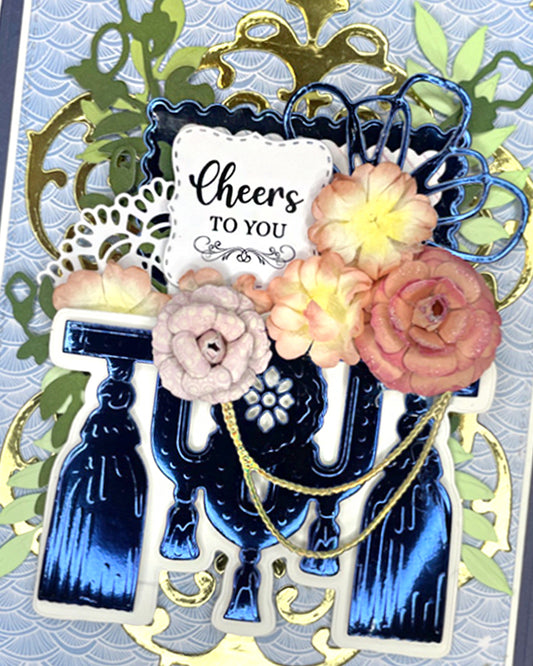 Close-up of a handmade luxury card showing layered blush, peach and dusty rose paper roses with green foliage, a gold ornate filigree frame, and a white sentiment tag reading "Cheers To You" in script. Navy and gold typographic elements visible below. Handmade greeting card made in New Zealand.