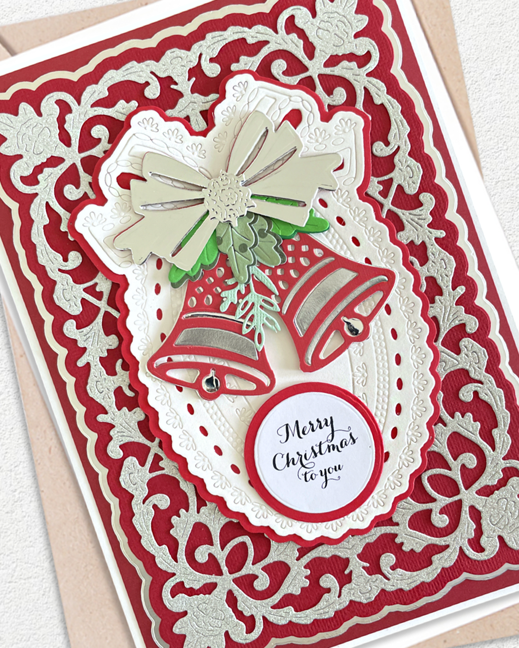 A close-up of a handmade Christmas greeting card featuring layered red and white scalloped paper borders, two red bells with green holly accents, a white bow, and a round white label that reads 'Merry Christmas to you'.