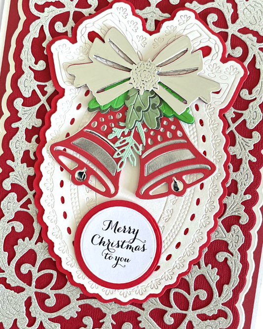 A close-up of a handmade Christmas greeting card featuring two red and silver bells, a white bow, green leaves, and a round tag that reads 'Merry Christmas to you' on a red and white patterned background.