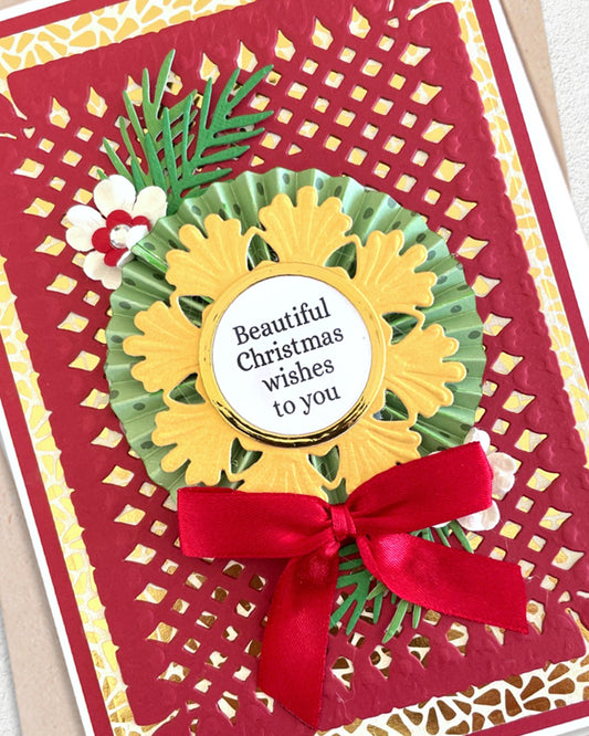 A close-up of a handmade Christmas greeting card featuring a red and gold laser-cut background, a yellow and green paper sunflower, a red ribbon bow, and a white circular label that reads 'Beautiful Christmas wishes to you'.
