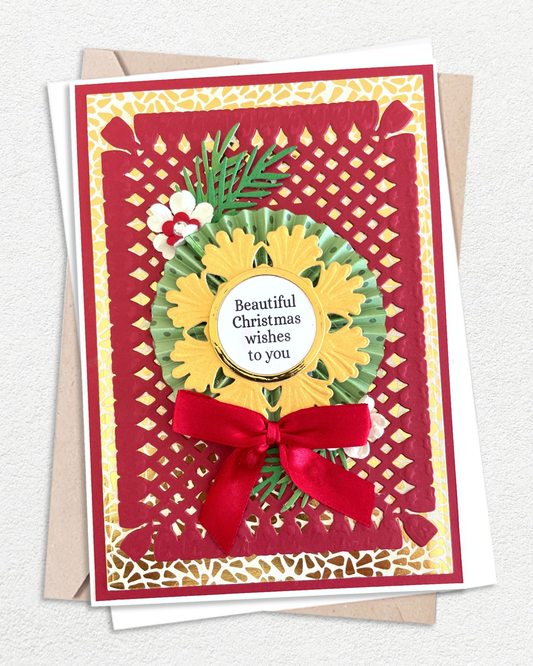 A handmade Christmas greeting card with a red and gold lattice border, a yellow sunflower, green leaves, and a red ribbon bow, with a circular label reading 'Beautiful Christmas wishes to you.' It is shown with kraft envelopes on a light textured background. Made by Del Cards Nz.