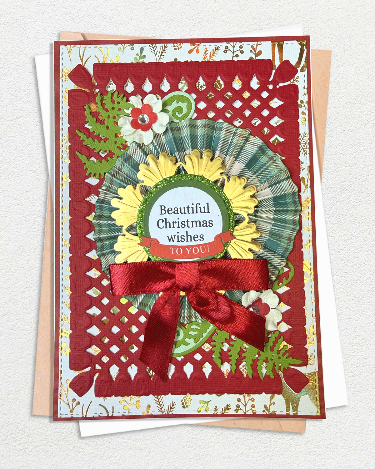 A handmade layered Christmas greeting card with a red lattice border, a green and yellow wreath, red ribbon bow, and floral details, displayed on top of a kraft envelope and white card against a textured off-white background.
