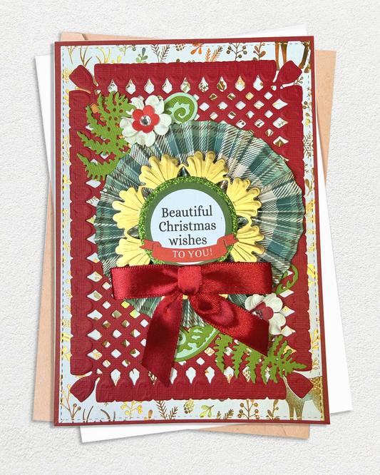 A handmade layered Christmas greeting card with a red lattice border, a green and yellow wreath, red ribbon bow, and floral details, displayed on top of a kraft envelope and white card against a textured off-white background.