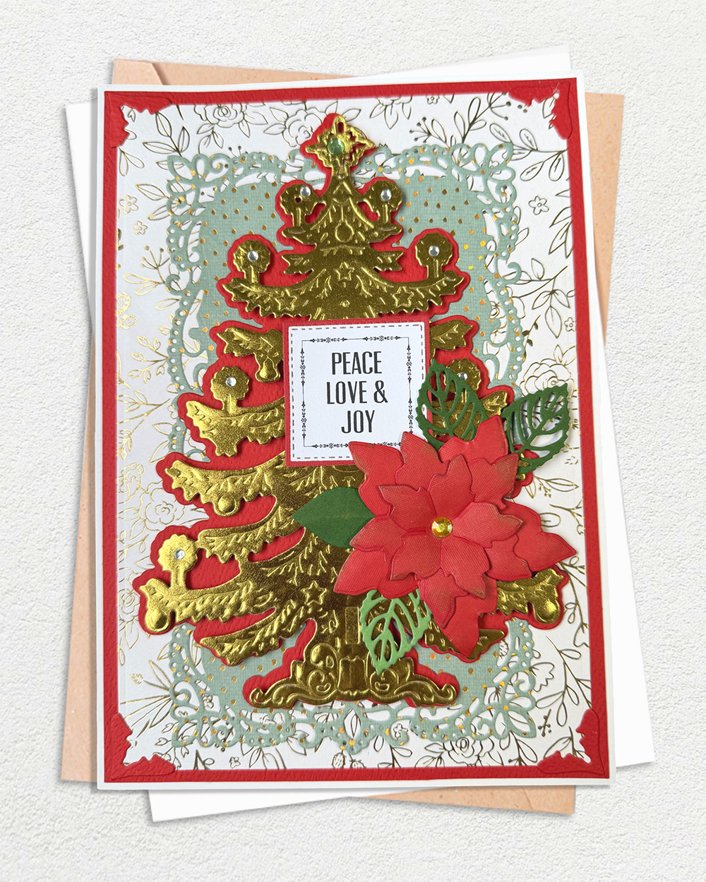 A handmade Christmas greeting card featuring a layered gold Christmas tree, a red poinsettia flower, green leaves, and a central white label that reads 'PEACE LOVE & JOY'. The card is styled on top of a kraft envelope against a textured off-white background.