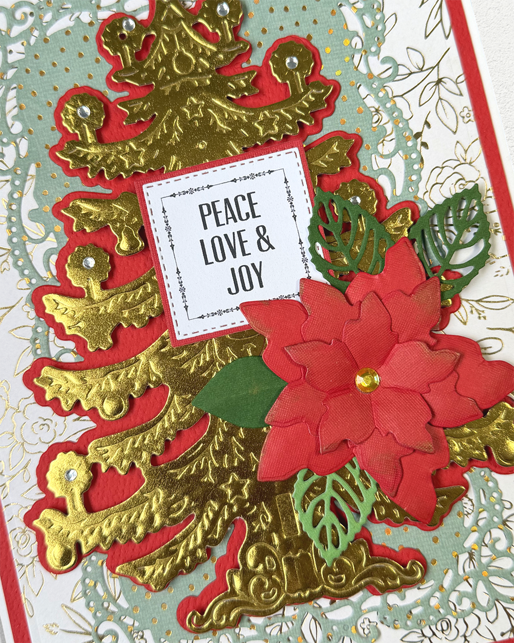 A close-up of a handmade holiday card featuring layered red and gold die-cut paper designs, including a Christmas tree, poinsettia flower, and a small white card reading 'PEACE LOVE & JOY'.