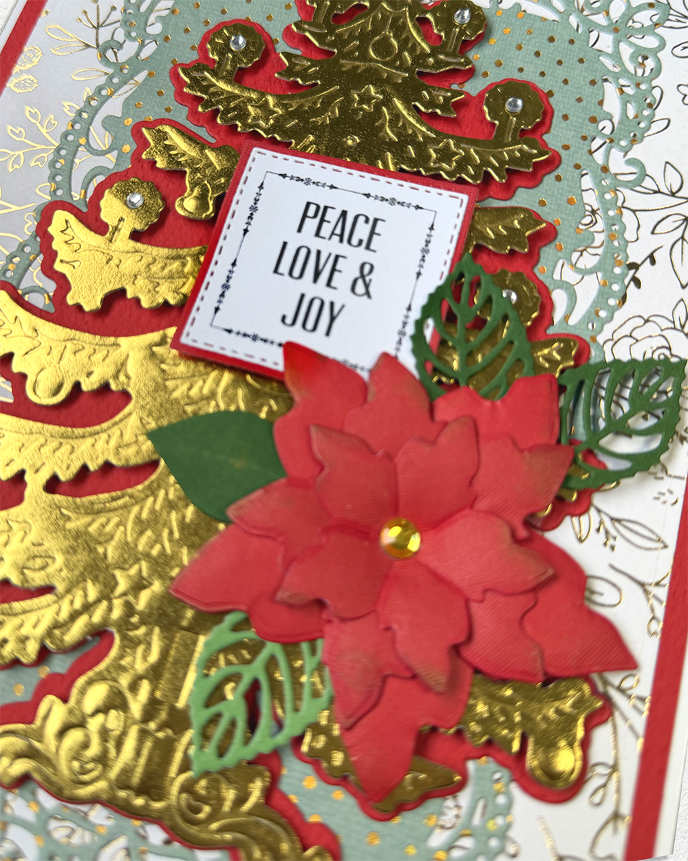 A close-up of a handmade Christmas greeting card featuring a red poinsettia flower, green leaf cutouts, gold foil ornaments, and a white paper label that reads 'PEACE LOVE & JOY'.