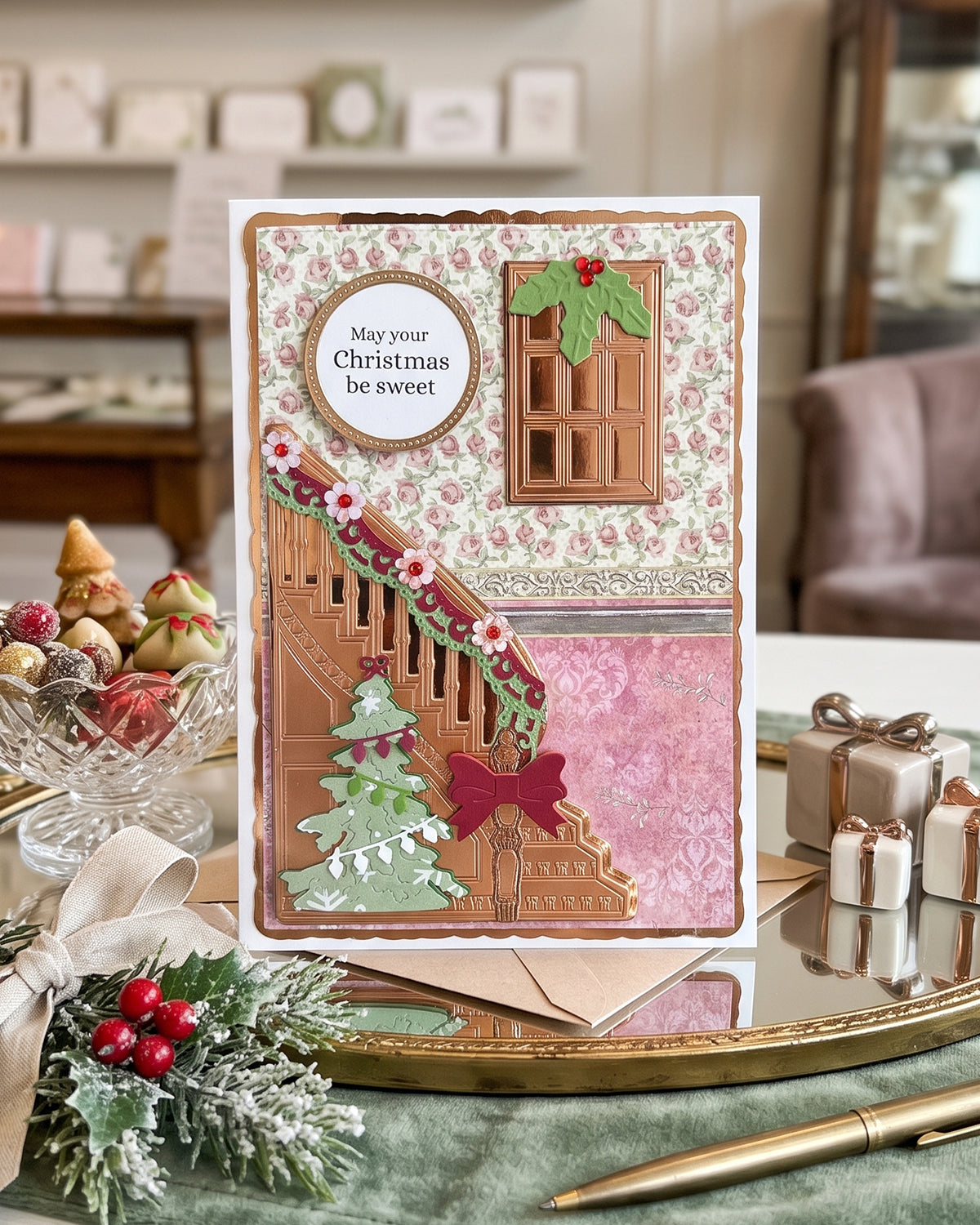 A handmade Christmas greeting card displayed on a tabletop in a decorated holiday setting. The card features a scalloped border, a floral patterned background, a brown staircase with a green Christmas tree and red bow, a window with holly, and a circular label that reads, 'May your Christmas be sweet'. Made in New Zealand by Del Cards NZ.