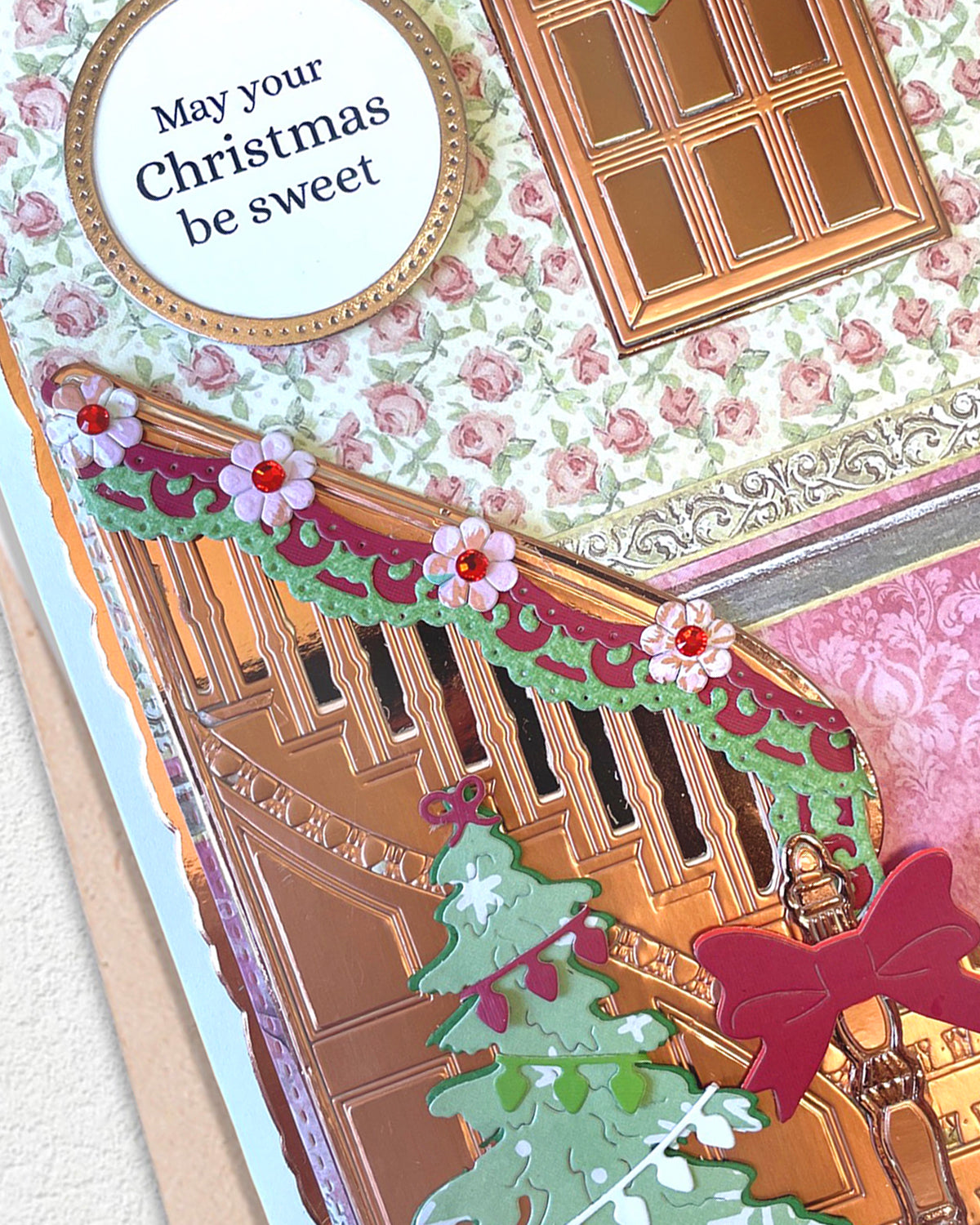 A close-up of a handmade Christmas card featuring a brown wooden door, a green garland with pink flowers, a decorated Christmas tree, a red bow, and a round white label that reads 'May your Christmas be sweet'.
