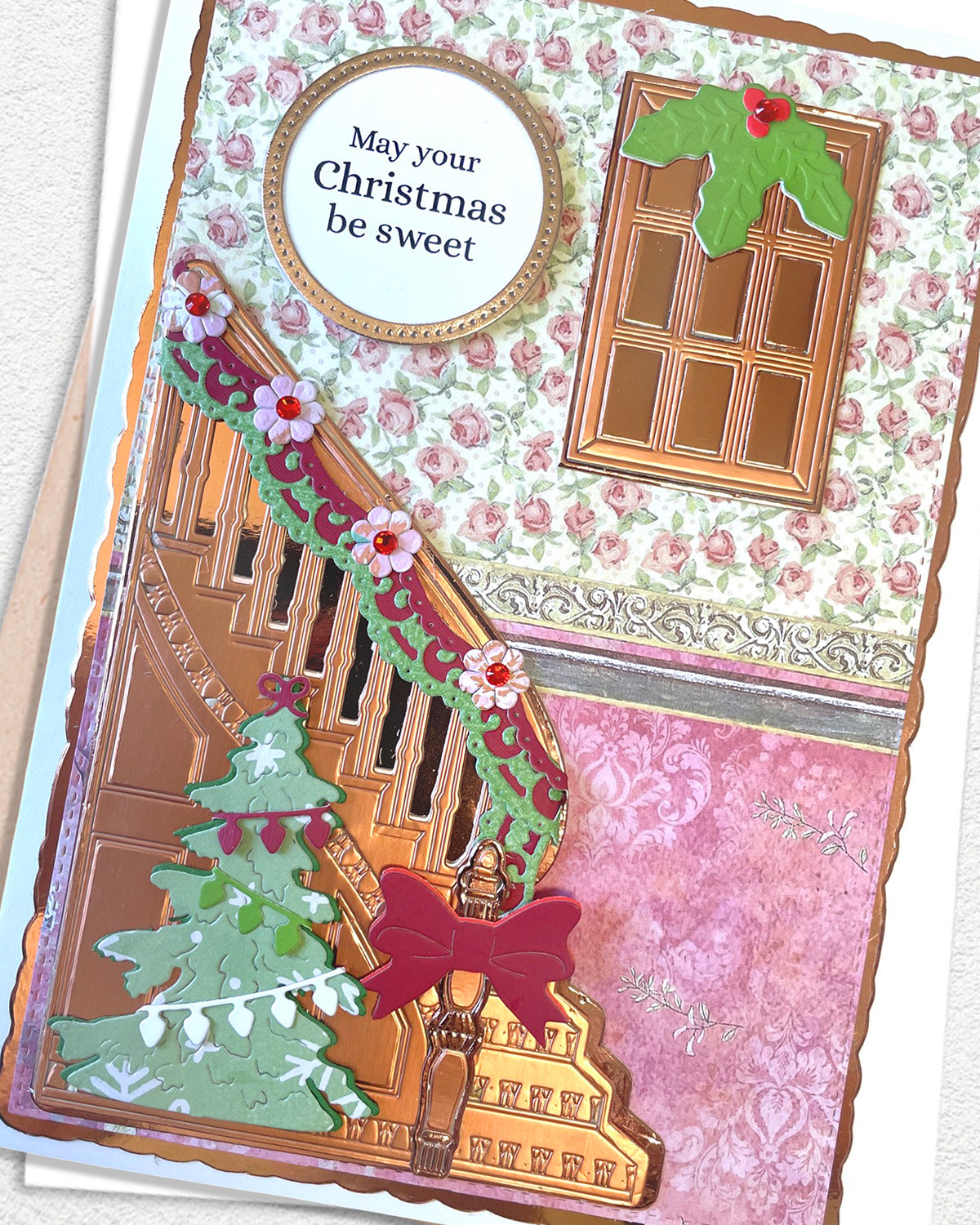 A close-up of a handmade Christmas greeting card featuring a decorated staircase, a green Christmas tree, a pink floral background, a window with holly, and a round label that reads 'May your Christmas be sweet'.