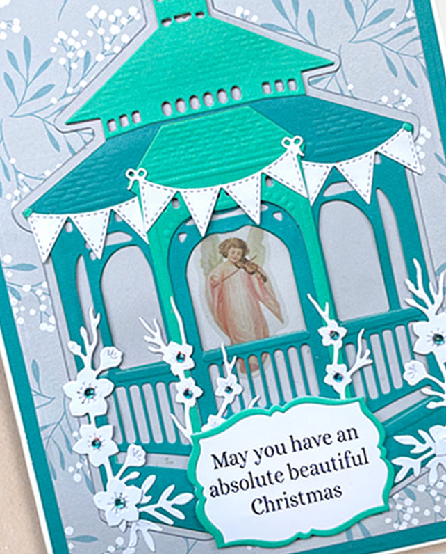 A close-up of a handmade layered Christmas card featuring a teal and white pagoda design with a small angel illustration in the center and a white banner that reads, 'May you have an absolute beautiful Christmas.'.