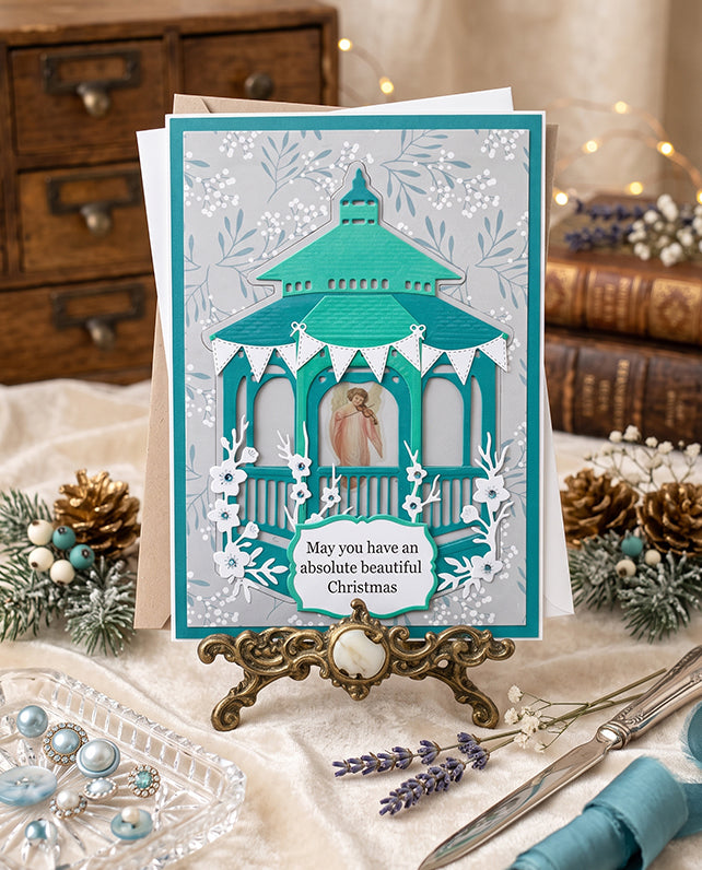 A handmade Christmas greeting card displayed on an ornate gold stand. The card features a layered paper design of a teal gazebo with white snowflakes and the text 'May you have an absolute beautiful Christmas'.