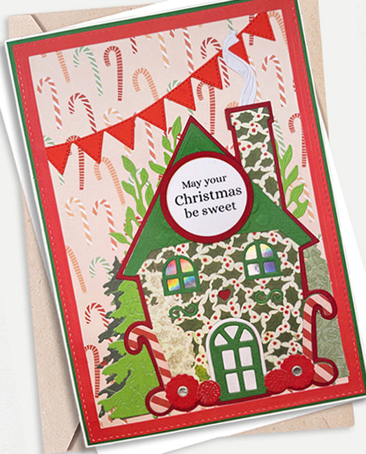 A handmade Christmas greeting card featuring a decorated gingerbread house, candy canes, greenery, and red banner flags, with the text 'May your Christmas be sweet.' It is shown layered over a kraft envelope on a light neutral background.
