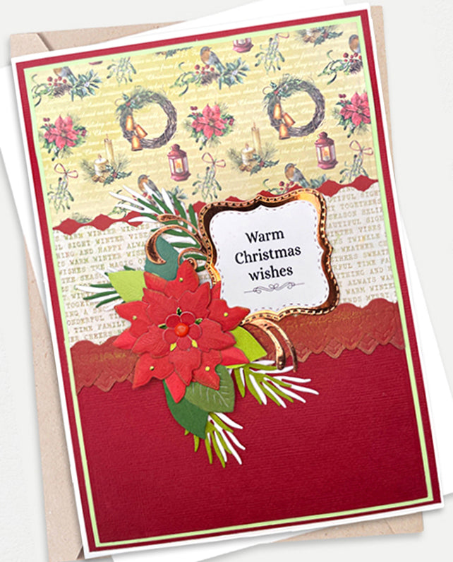 A handmade Christmas greeting card with a red poinsettia, green foliage, and a central label reading 'Warm Christmas wishes', shown with a kraft envelope on a white background.