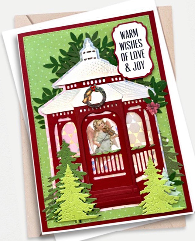 A festive holiday greeting card featuring a red gazebo with an angel playing a violin, green trees, and a light green polka-dot background. The card reads 'WARM WISHES OF LOVE & JOY' and is shown with a kraft envelope behind it.