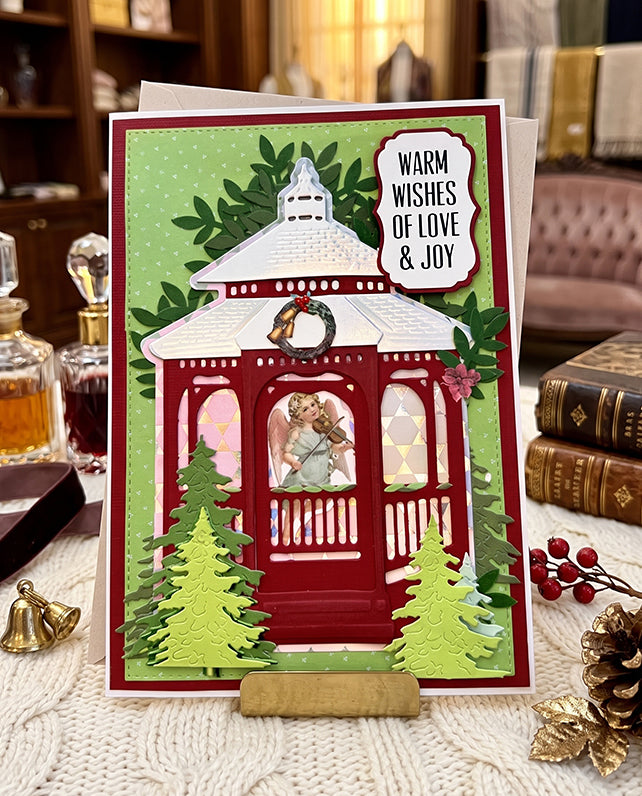 A handmade Christmas greeting card displayed on a small wooden stand on a knitted table runner. The card features a red gazebo with an angel figure inside, green trees, a wreath, and a green polka-dot background.